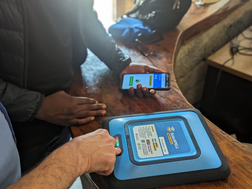 Close-up of a man's hand pressing a button on the SolarSPELL library hardware and another man holding a smartphone displaying a module in the SolarSPELL library.